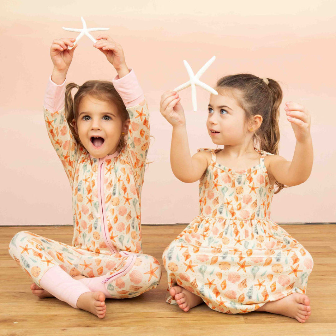 Two girls sitting down in our Sandy Seashell Collection. One is wearing our convertible bamboo pajama and the other one our Sundress. 