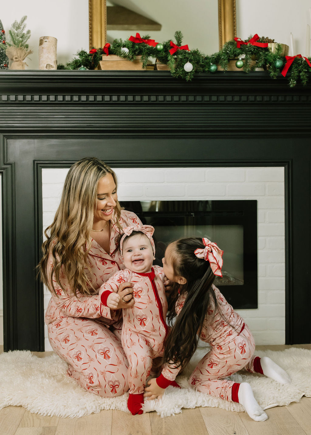Close-up of girls wearing Candy Cane Coquette Bamboo Bow Hair Clips with coordinating pink and red holiday outfits beside a decorated fireplace.