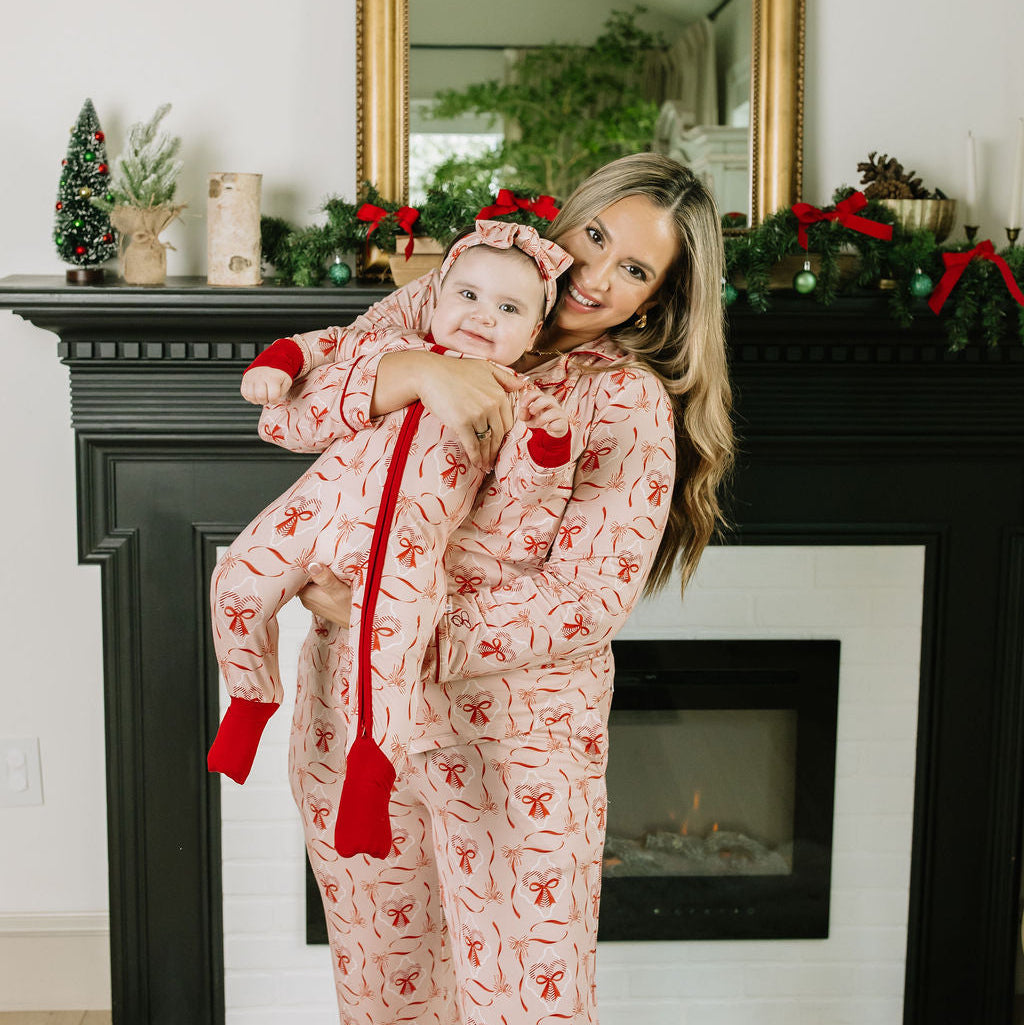 Mother holding baby wearing Candy Cane Coquette Bamboo Convertible Footie Romper in front of a decorated holiday fireplace.