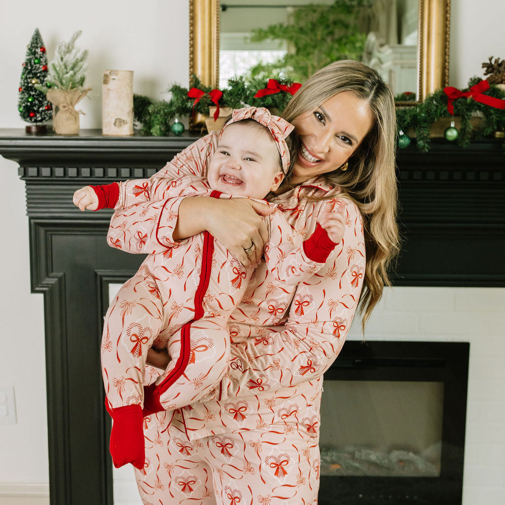 Mother smiling with baby in matching Candy Cane Coquette holiday bamboo pajamas beside a fireplace.