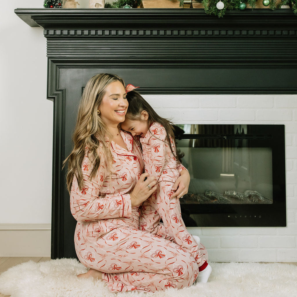 Mother and daughter wearing matching Candy Cane Coquette bamboo pajamas in a festive pink and red holiday print, sitting together in front of a fireplace.