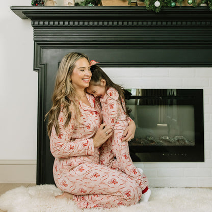 Mother and daughter wearing matching Candy Cane Coquette bamboo pajamas in a festive pink and red holiday print, sitting together in front of a fireplace.