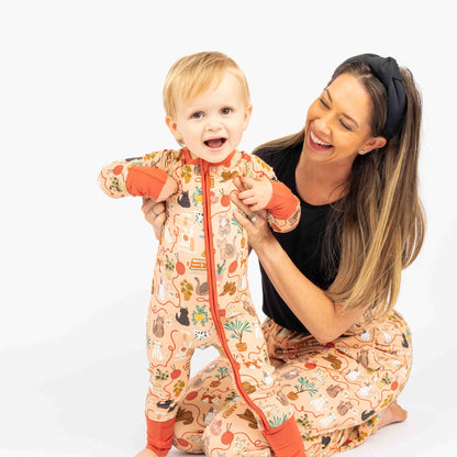 Mom and baby smiling  and sitting while modeling our pet cat print that is orange with red/orange trim. Features cats, plants, books and yarn on the bamboo pajamas as well.