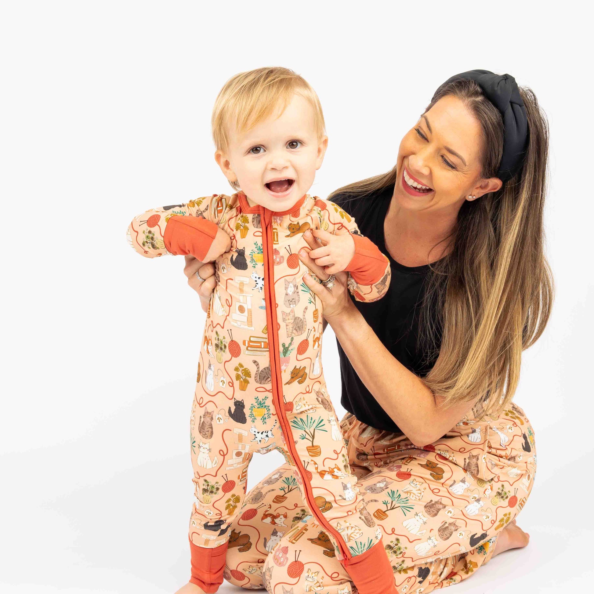 Mom and baby smiling  and sitting while modeling our pet cat print that is orange with red/orange trim. Features cats, plants, books and yarn on the bamboo pajamas as well.