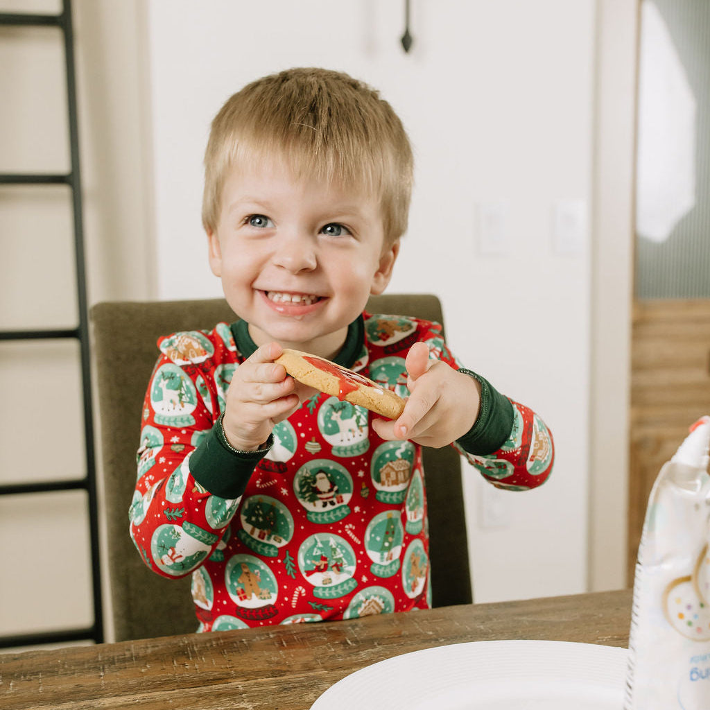 Smiling toddler boy wearing festive Christmas bamboo pajamas while sitting at a wooden table.