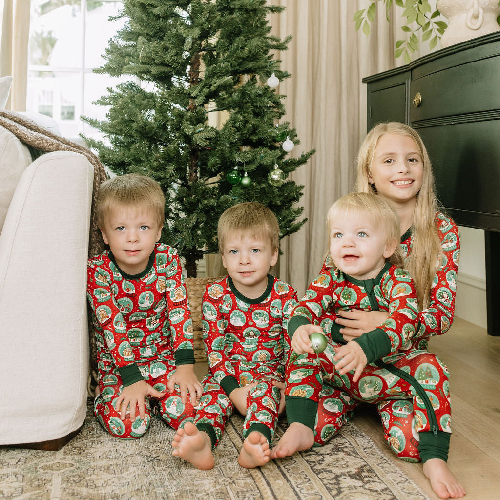 Three children wearing matching red and green Christmas bamboo pajamas while sitting beside a decorated Christmas tree.