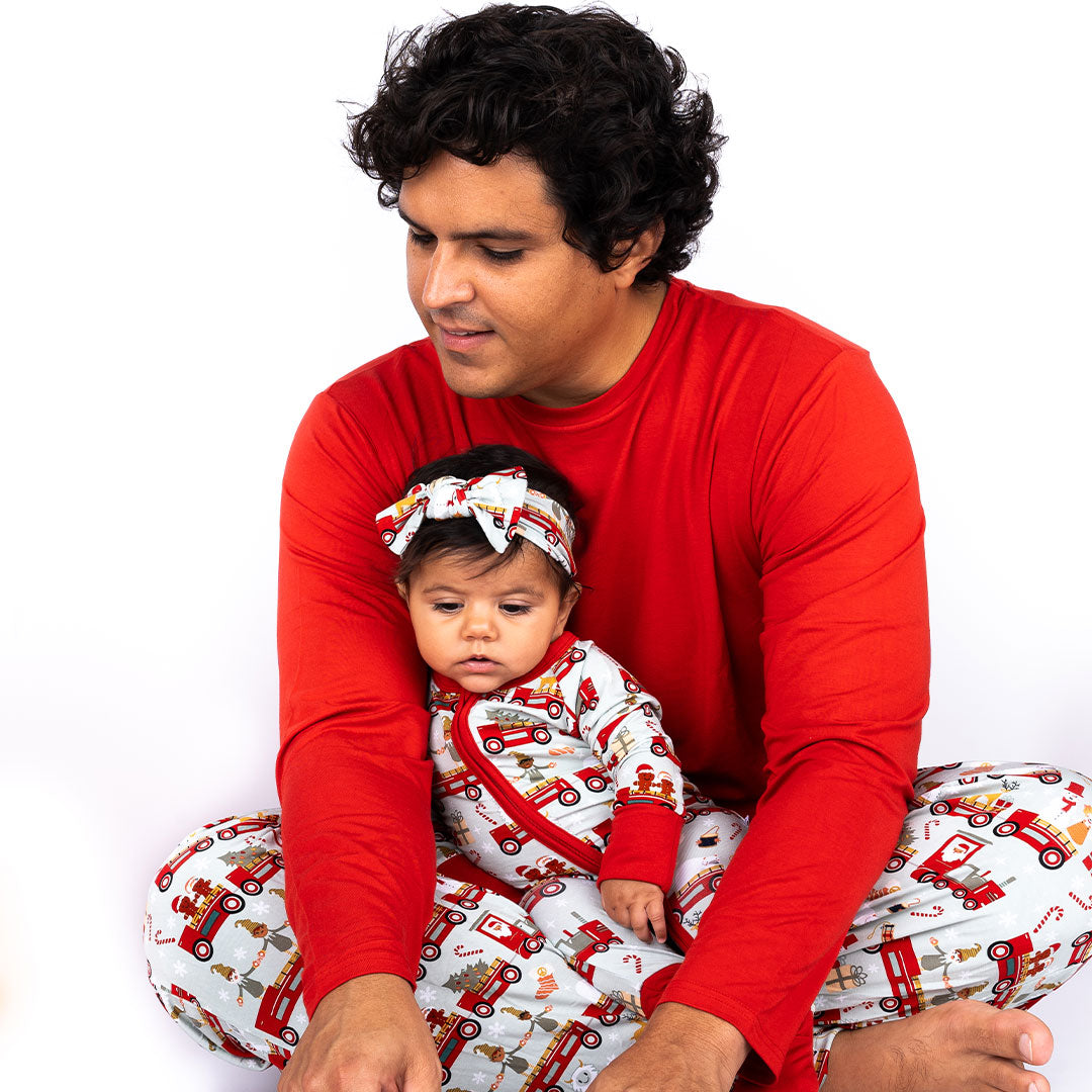 father sits criss-cross and holds his baby in his lap. the baby wears the "christmas train" baby headband and convertible. the father wears the relaxed pants. the "christmas train" pattern has various winter character that stand/ride on a red train. these characters include: santa, santa's elves, raindeers, yeti monsters, gingerbreads, reindeer, christmas trees, candy canes, presents, and snowflakes. 