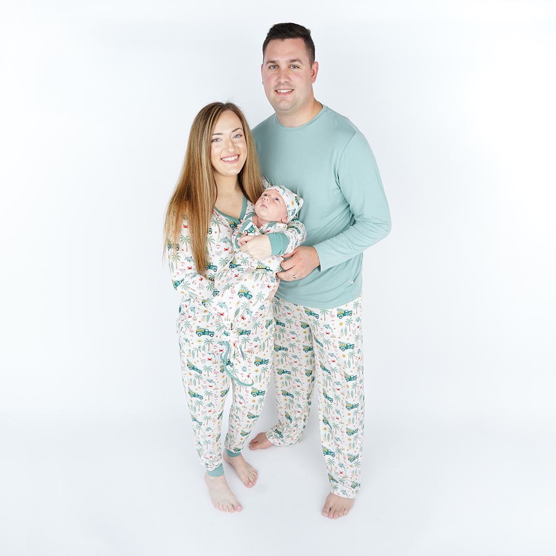 family of 3 smile and pose for the camera. the mother stands on the left in the "coastal christmas" women's top and jogger pants. she holds her infant who is in the "coastal christmas" gown and hat set. the father, who stands on the right, is in the "coastal christmas" relaxed pants. the "coastal Christmas" print is a mix of sandman, beach Santas, decorated palm trees, flamingos with Santa hats, dolphins with reindeer antlers, craps, suns, shells, and starfish scattered around a white background. 