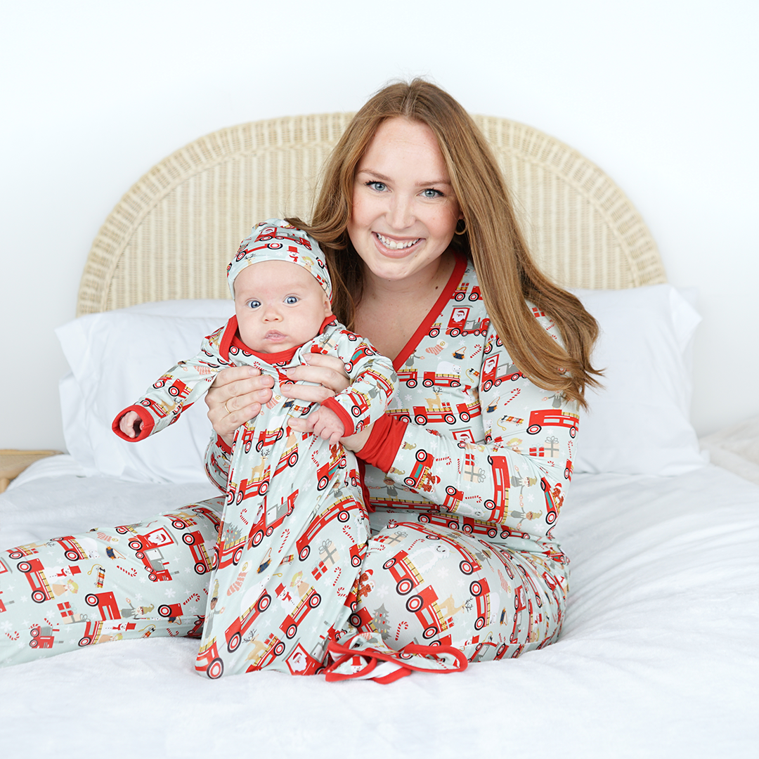 mom sits on a bed and holds up her baby. they are both wearing "christmas train" apparel. the mom in the women's top and the jogger bottoms. the baby is in the gown and hat set. the "christmas train" pattern has various winter character that stand/ride on a red train. these characters include: santa, santa's elves, raindeers, yeti monsters, gingerbreads, reindeer, christmas trees, candy canes, presents, and snowflakes. 