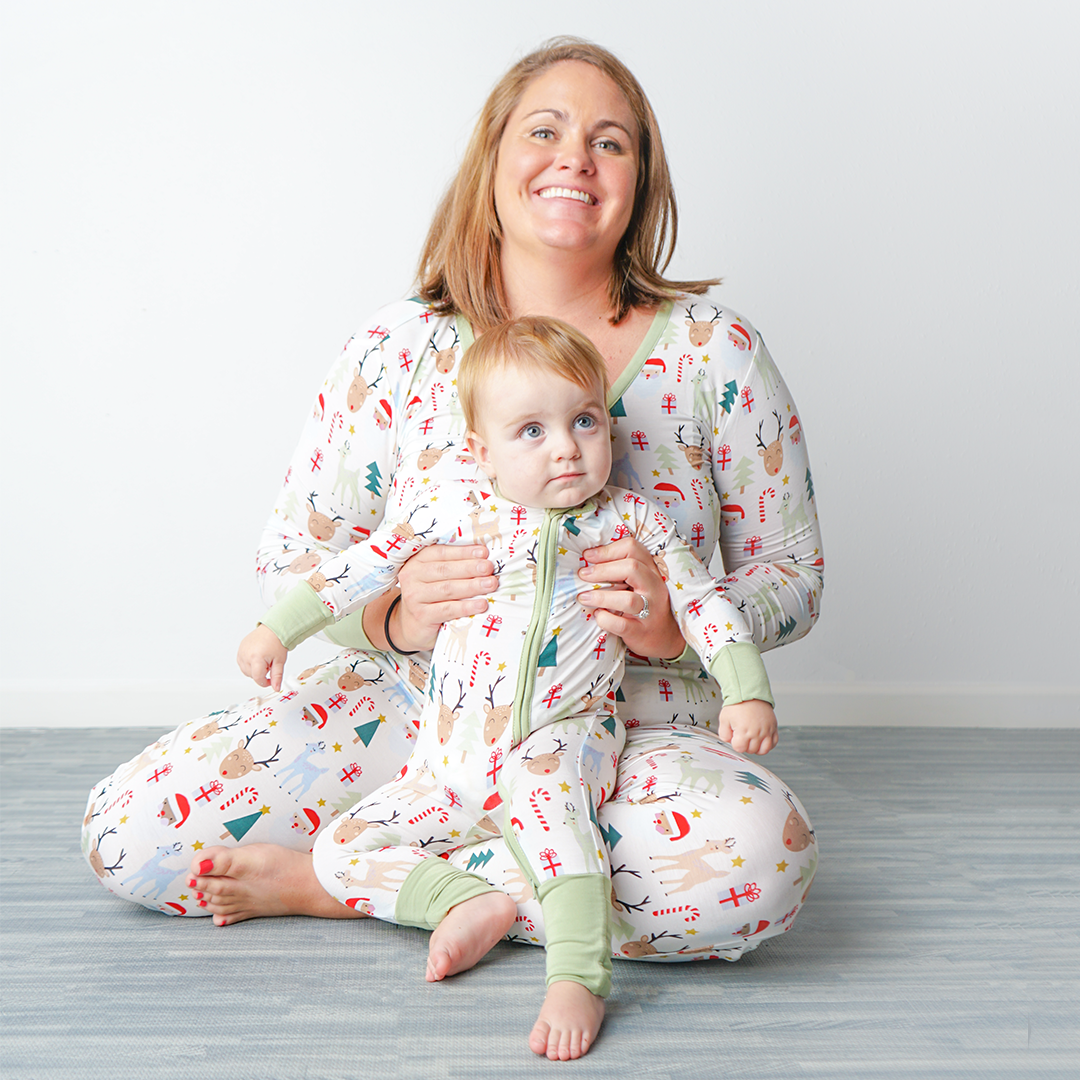 woman sits on the floor and hold up her baby. they are both in "Santa and friends" attire. mom wears the jogger pants and women's top. the baby is in the convertible. the "Santa and friends" print is a Christmas themed pattern mixed with Santa heads, candy canes, reindeer, Christmas trees, Rudolph heads, and gold stars. this is all on a white background. 