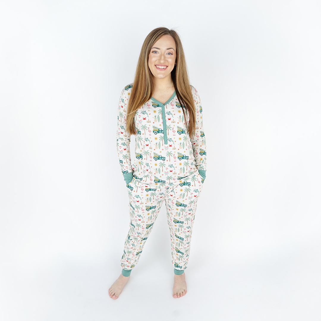 woman poses in the "coastal christmas" women's top and jogger pants. the "coastal Christmas" print is a mix of sandman, beach Santas, decorated palm trees, flamingos with Santa hats, dolphins with reindeer antlers, craps, suns, shells, and starfish scattered around a white background. 