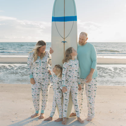 Family of four standing on a beach with a surfboard, all wearing matching pajamas.