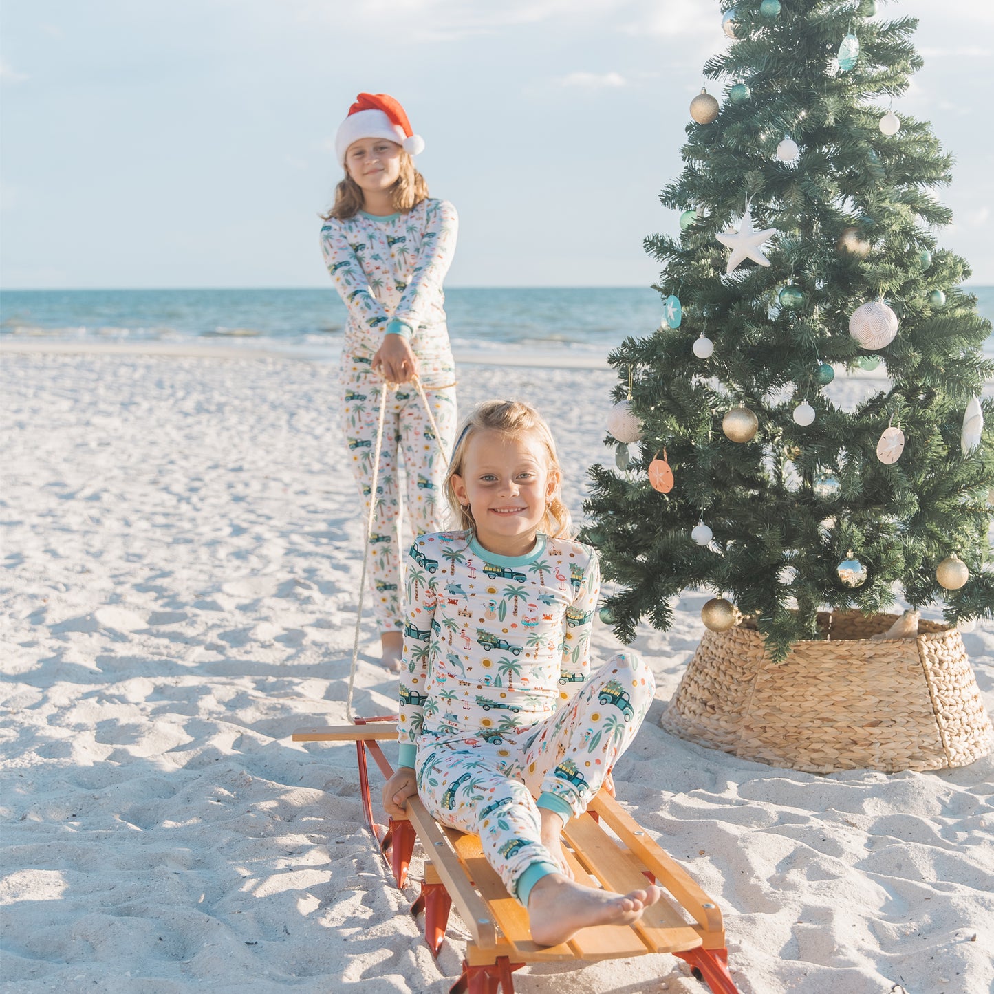 Two children in pajamas on a sled with a Christmas tree on a beach.