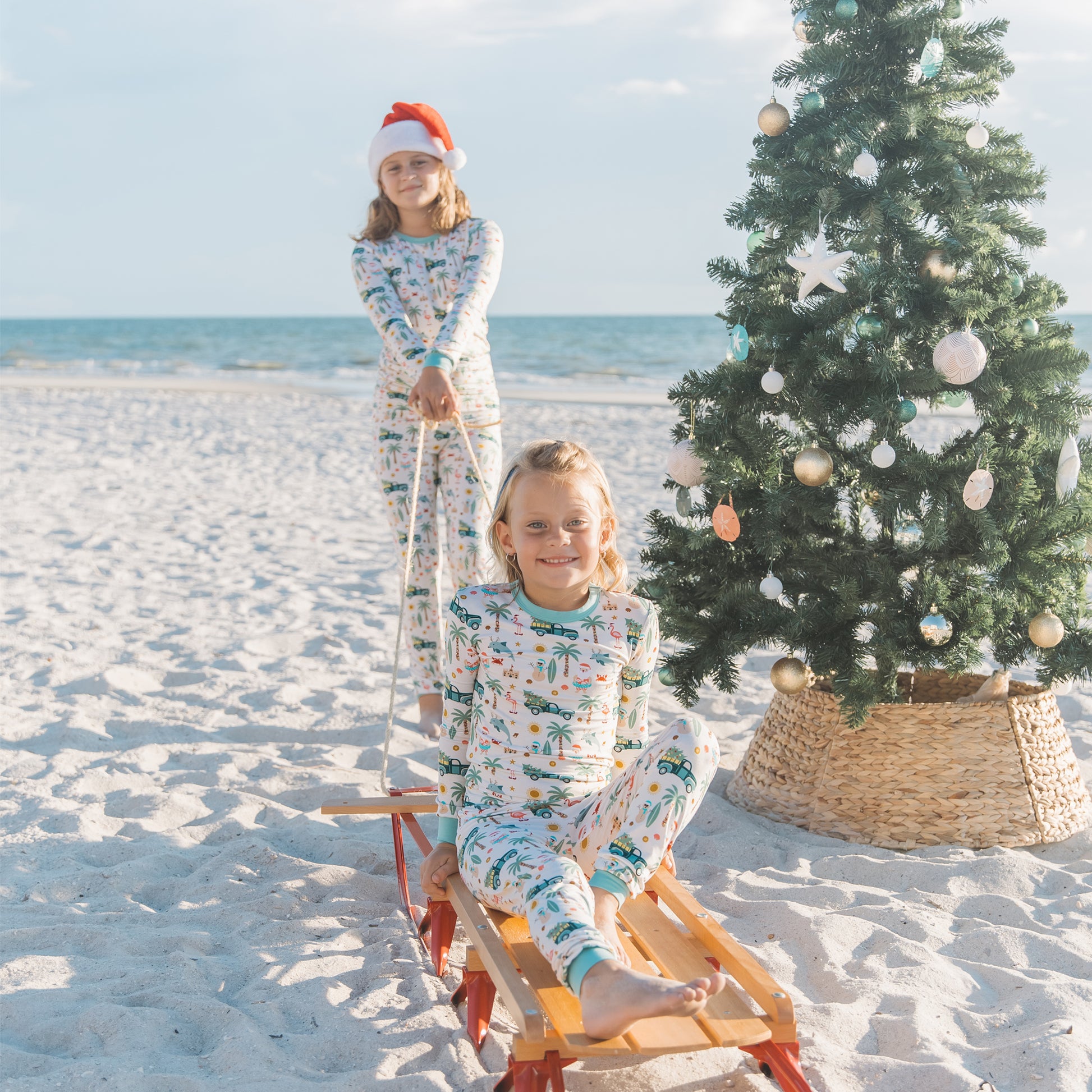 Two children in pajamas on a sled with a Christmas tree on a beach.