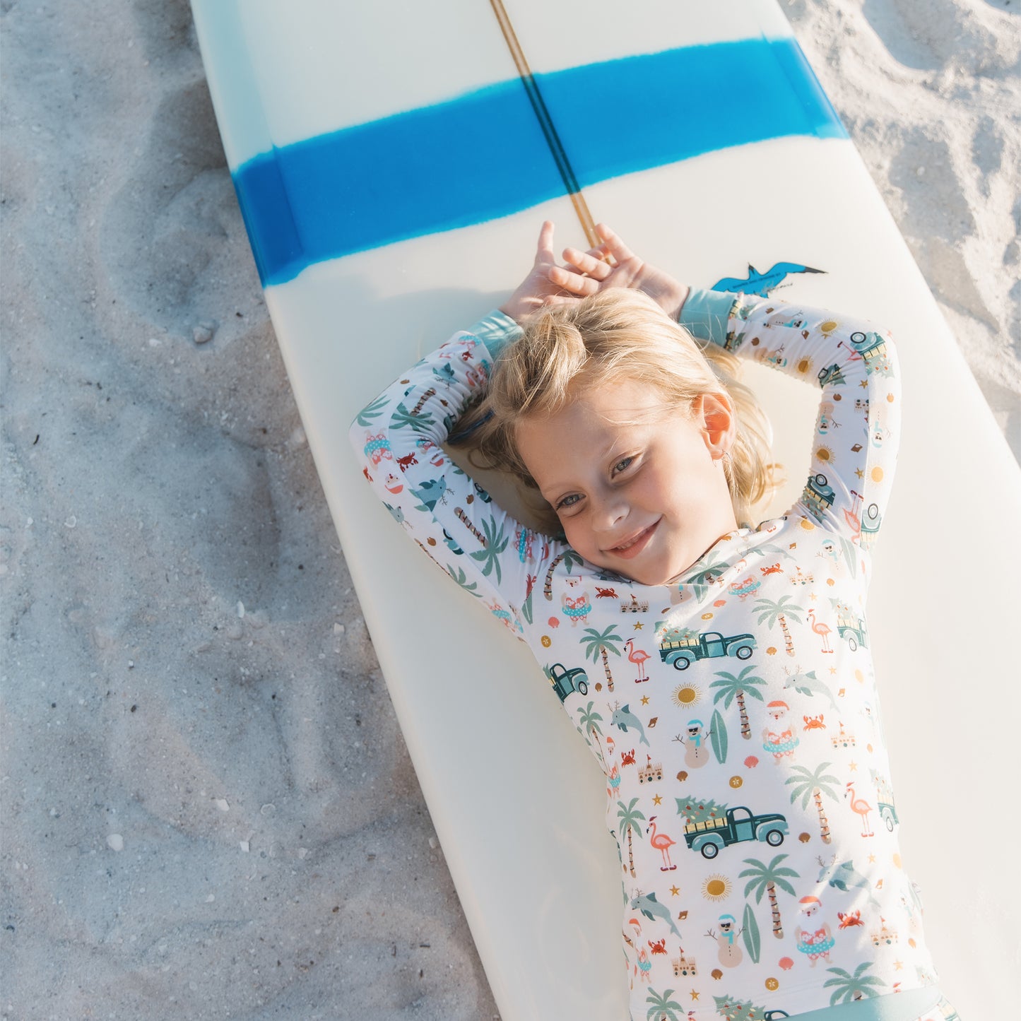 Child lying on a surfboard with a patterned shirt on a sandy beach