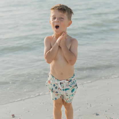 Toddler boy playing in the sand wearing Coral Reef Boys Swim Trunks, designed with a fun ocean-themed print of sharks and sea turtles, made from quick-dry, breathable fabric with UPF 50+ protection.