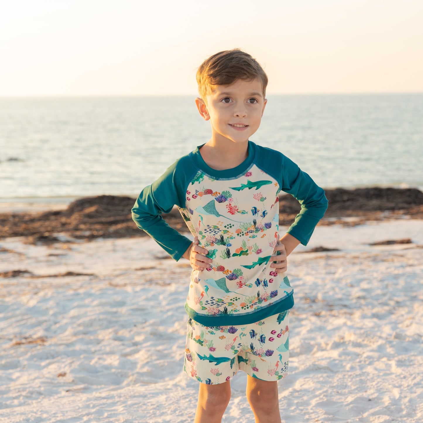 Young boy in a colorful swimsuit standing on a sandy beach with ocean in the background