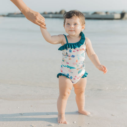 Toddler girl wearing a Coral Reef Ruffle Leg One Piece Swimsuit with an ocean-themed print of sharks and sea turtles, designed with soft, stretchy fabric and UPF 50+ sun protection for a fun day at the beach.