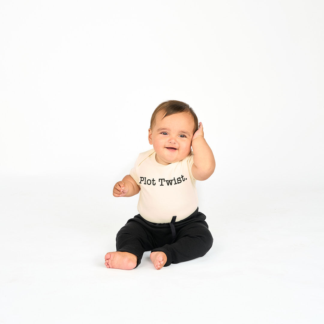 Baby Wearing white onsie with phrase "Plot Twist." and back cotton joggers while sitting and smiling at the camera