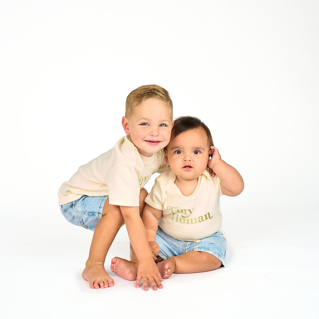 2 boys sitting and smiling at the camera while wearing white onesie/tees with the phrase "Tiny Human"