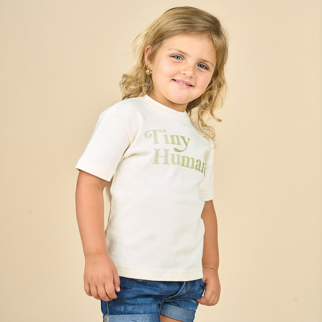 Girl smiling and looking into camera while waring jean shorts and a white cotton tee with the phrase "Tiny Human" 