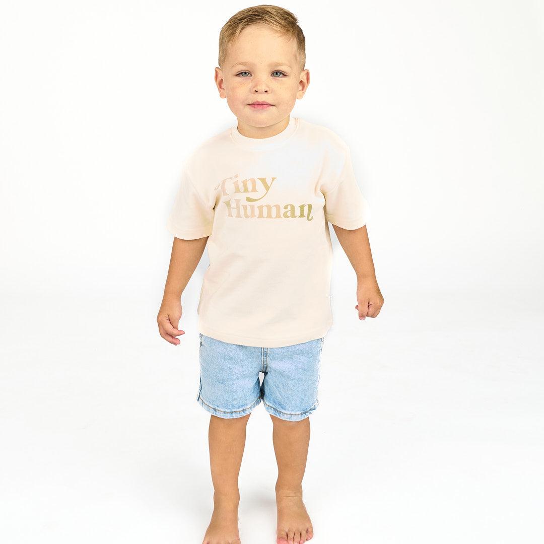 Boy looking at camera while wearing jean shorts and a white cotton tee with the phrase "Tiny Human"