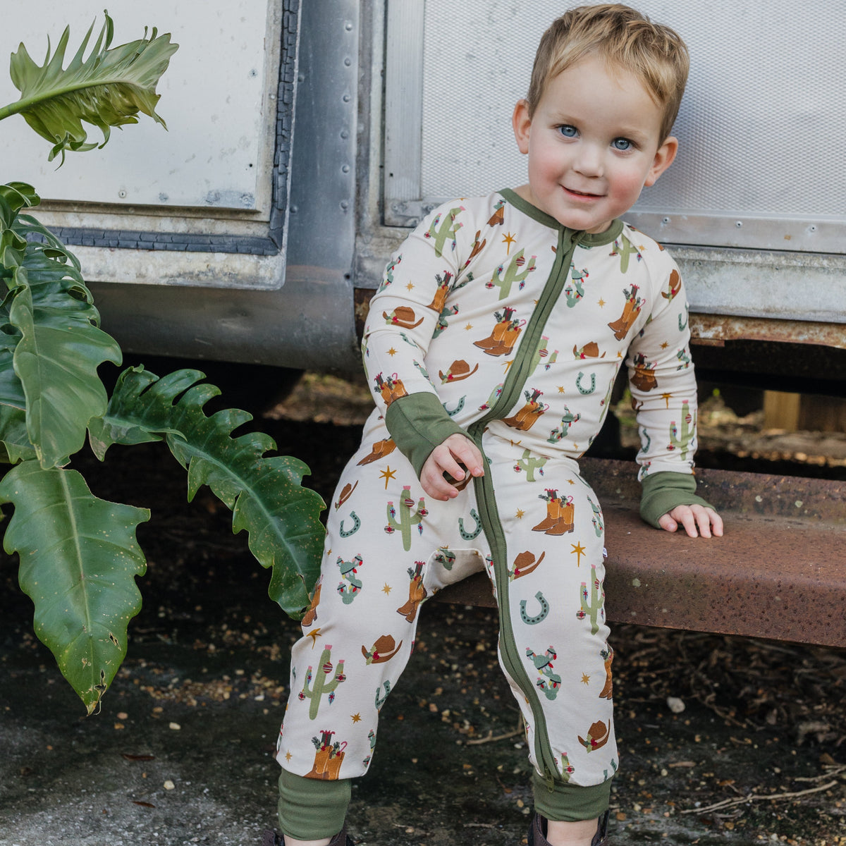 Toddler boy wearing Cowboy Christmas Bamboo Convertible Footie Romper with festive Western holiday print and green cuffs while sitting on wooden steps outdoors.