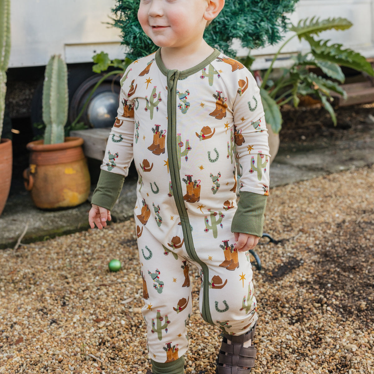 Toddler boy wearing Cowboy Christmas Bamboo Convertible Footie Romper with green cuffs and Western Christmas illustrations while standing outdoors.