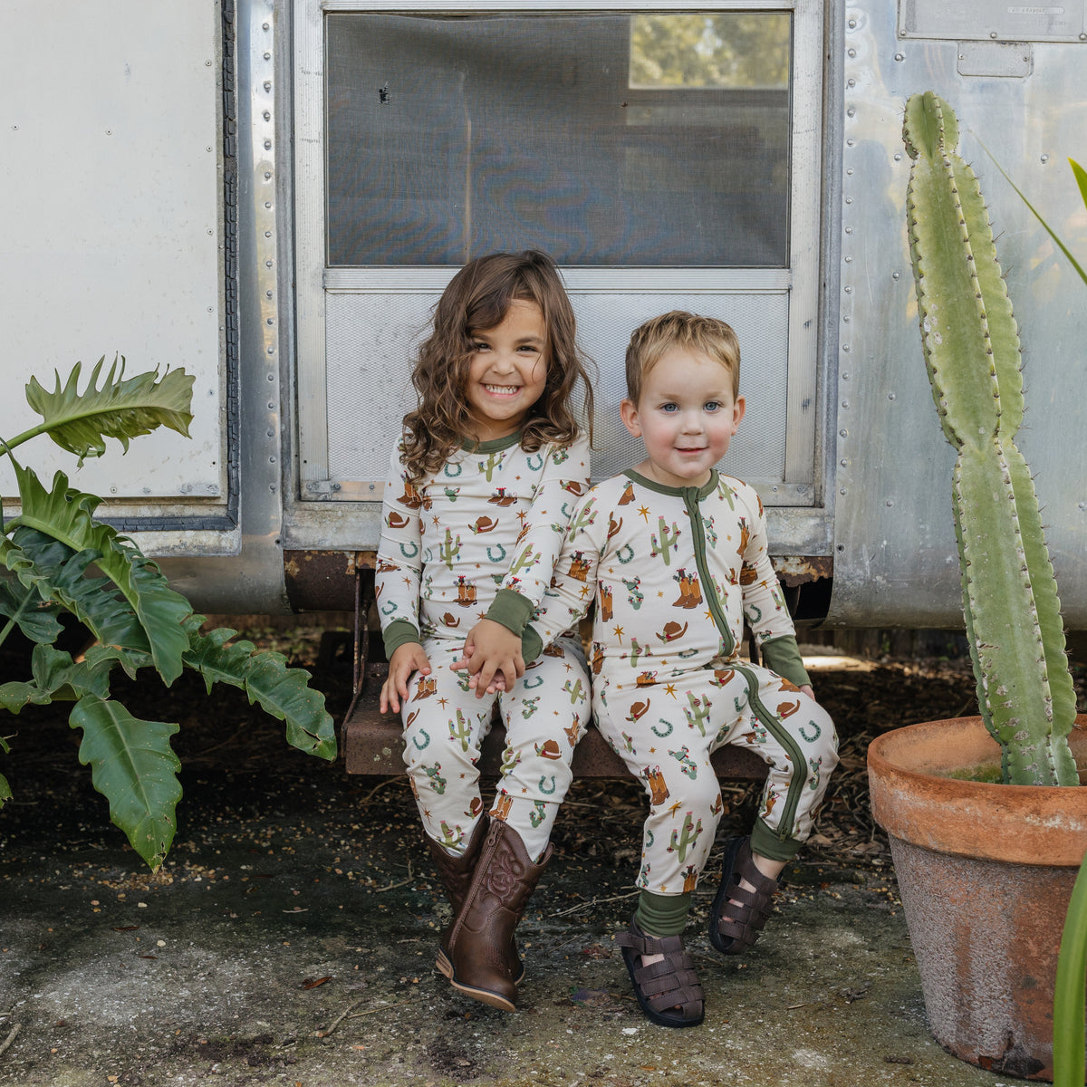 Two siblings wearing coordinating Cowboy Christmas bamboo outfits outdoors, one in a long sleeve two-piece pajama set and the other in a convertible footie romper, featuring festive Western holiday prints.