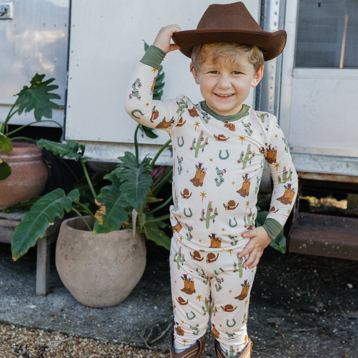 Toddler boy wearing Cowboy Christmas Bamboo Two-Piece Pajama Set with brown cowboy hat and boots while standing outdoors.