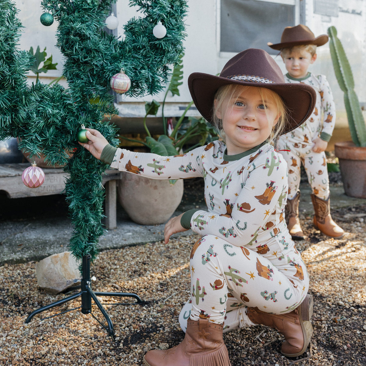 Smiling toddler boy wearing Cowboy Christmas Bamboo Two-Piece Pajama Set with cowboy hat and boots beside a decorated Christmas cactus.