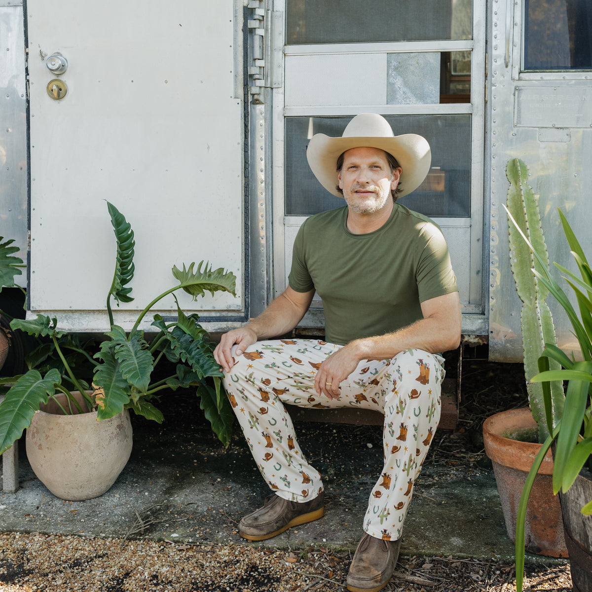 Man wearing Cowboy Christmas Bamboo Men's Lounge Pants with a green T-shirt and cowboy hat while sitting outside a vintage camper.