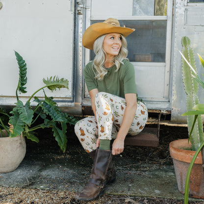 Woman wearing Cowboy Christmas Bamboo Joggers with a green shirt, cowboy hat, and boots while sitting on steps.