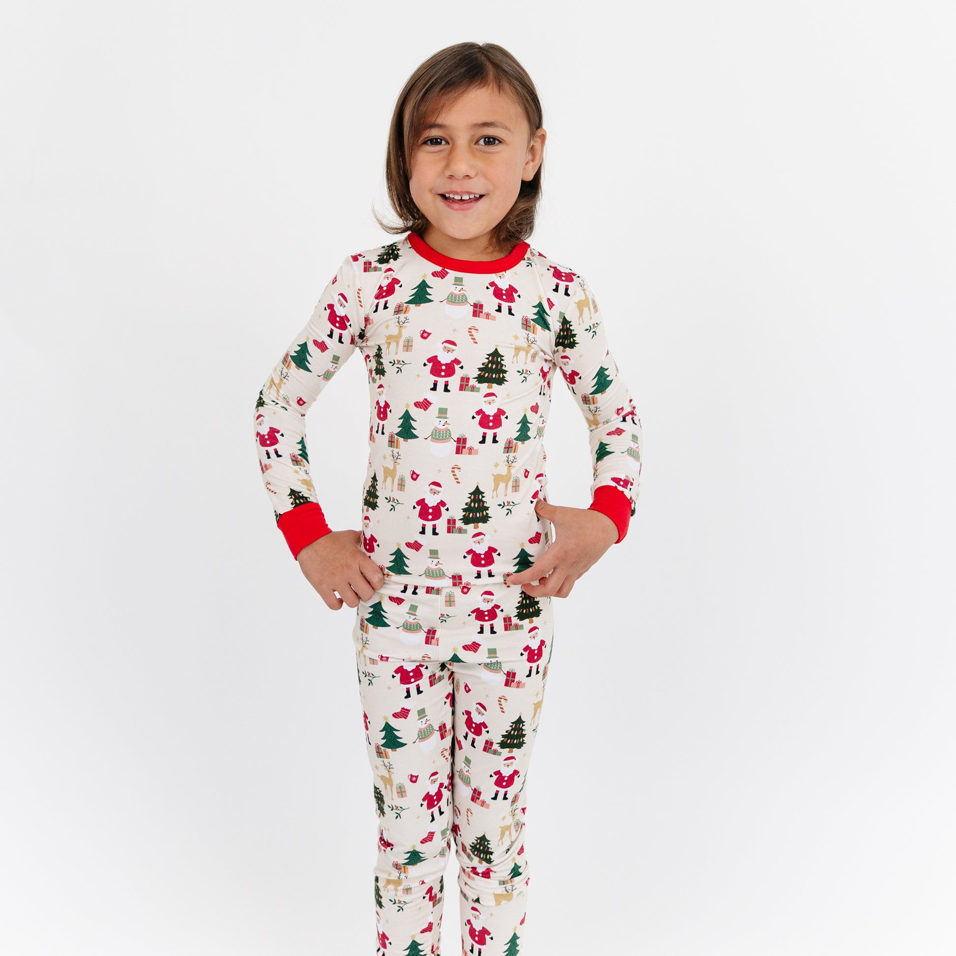 Child standing in studio wearing Cozy Christmas bamboo pajamas, featuring Santa and tree print with bright red cuffs.