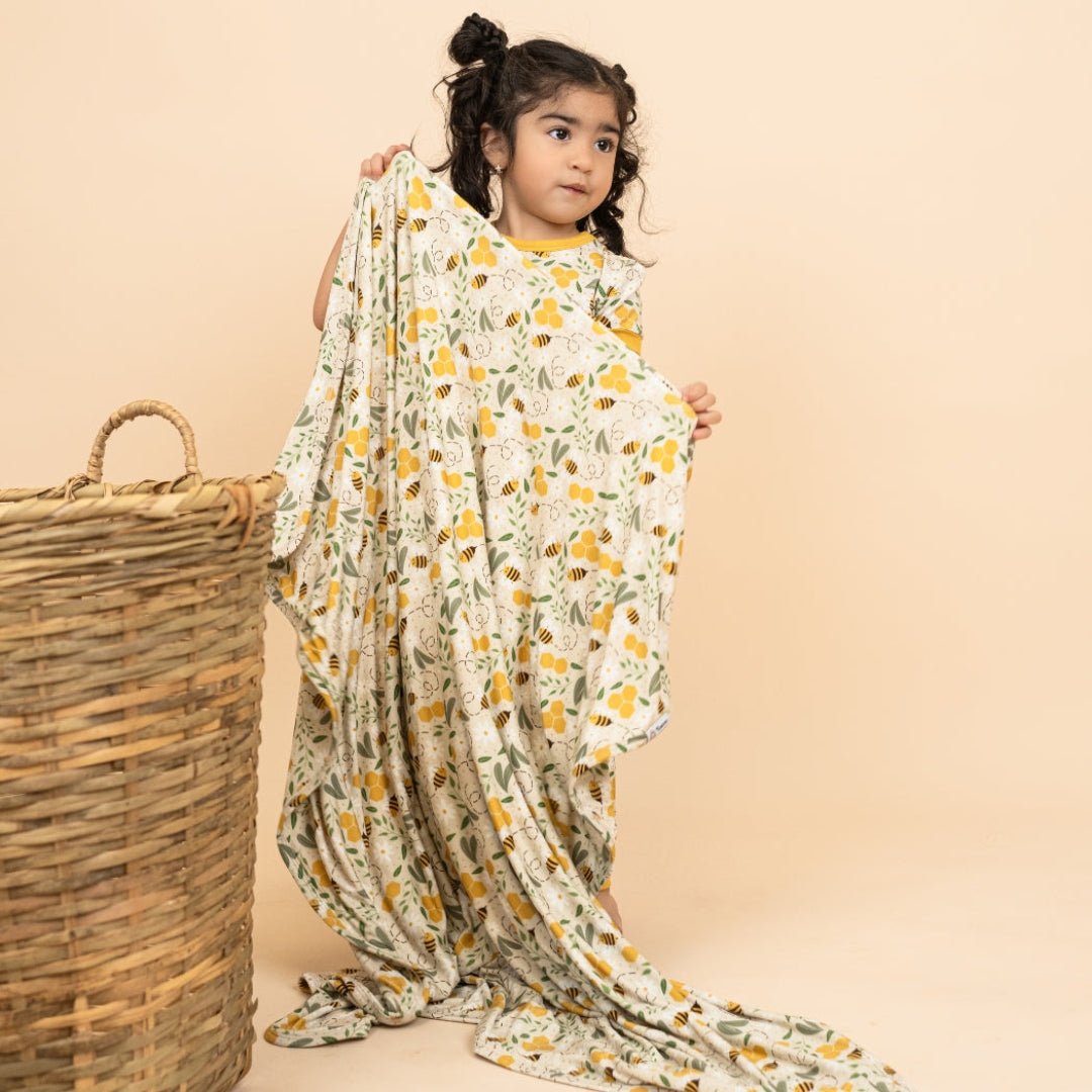 A young girl draped in the Happy Honey Bees luxury bamboo blanket by Emerson and Friends, featuring a charming honey bee and floral print. Made from ultra-soft, hypoallergenic bamboo fabric, this breathable blanket offers cozy comfort.