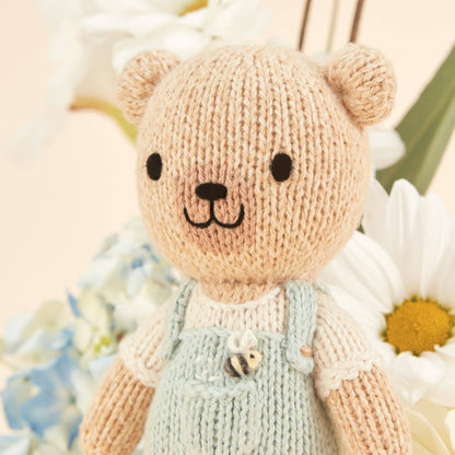 Knitted teddy bear with a light blue bib overalls sitting among flowers.