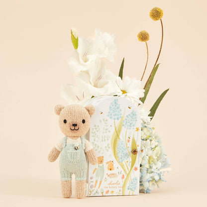 Knitted teddy bear in front of a floral arrangement and product packaging on a beige background