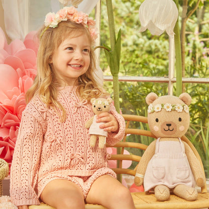 Young girl in a pink outfit holding a teddy bear with a floral headband, sitting next to a similar teddy bear in a garden setting.