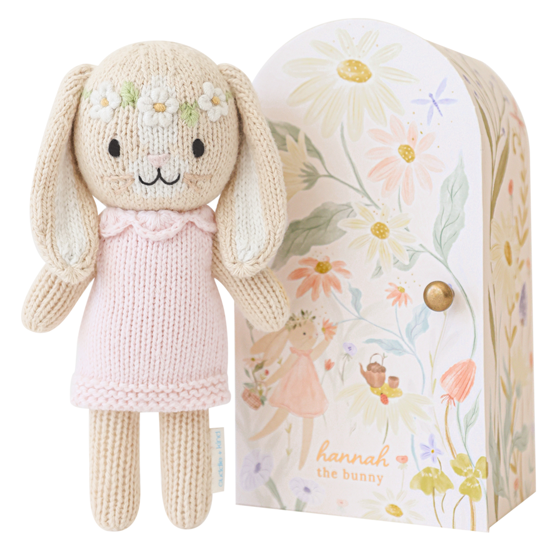 Knitted bunny doll with a pink dress in front of a floral-patterned box.