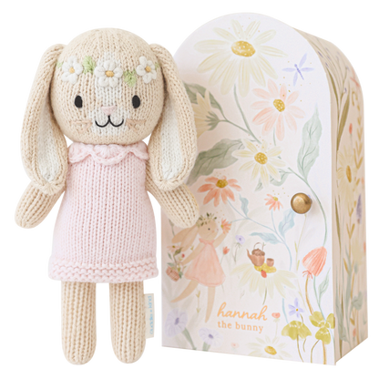 Knitted bunny doll with a pink dress in front of a floral-patterned box.