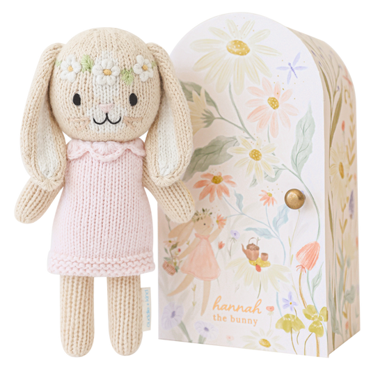 Knitted bunny doll with a pink dress in front of a floral-patterned box.