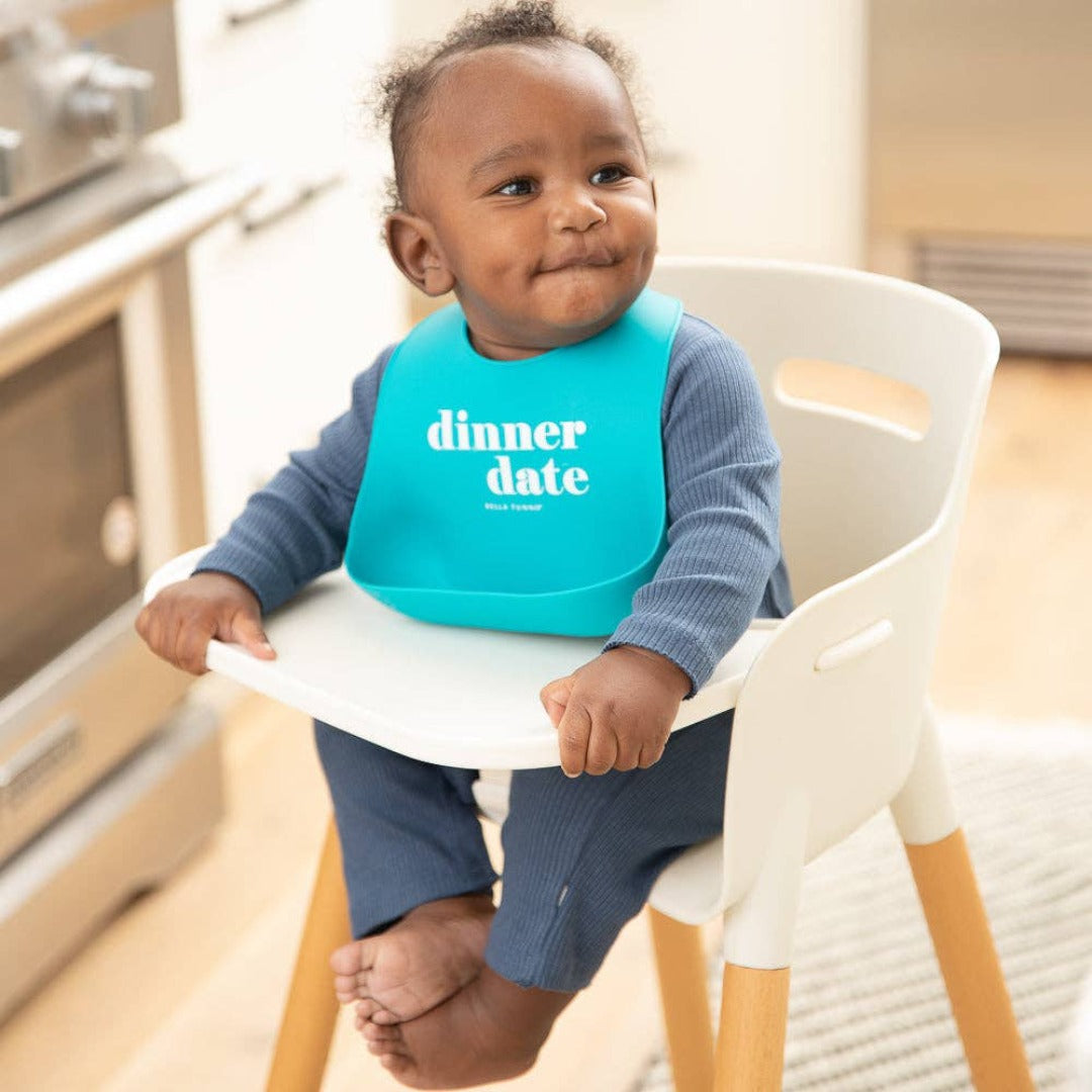 little boy sitting in a highchair wearing a blue bib that reads dinner date on the front in white