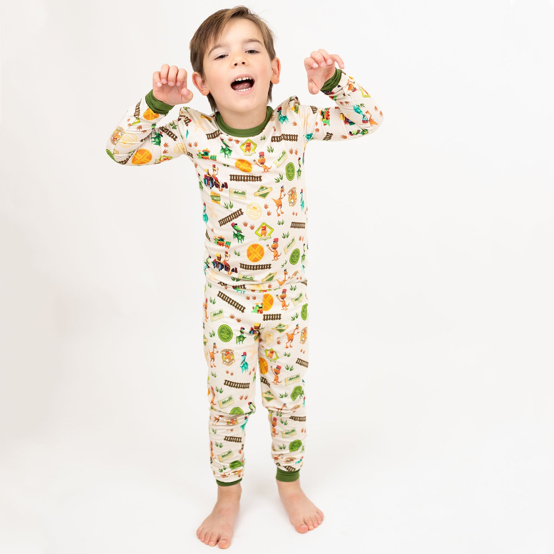 Toddler wearing our Emerson and Friends All Aboard the Dino Train Long Sleeve Kids Pajama Pants Set with green trim, and colorful orange, blue, and green dinosaurs on train tracks with "Dino Train" print.