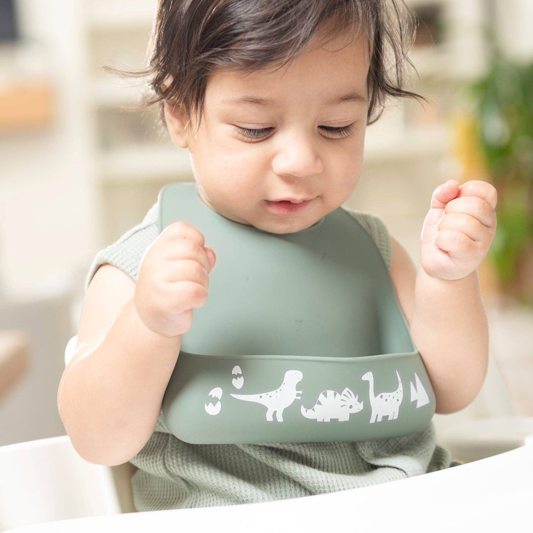 little boy in a highchair wearing a dark green silicone baby bib with white dinosaurs along the bottom edge