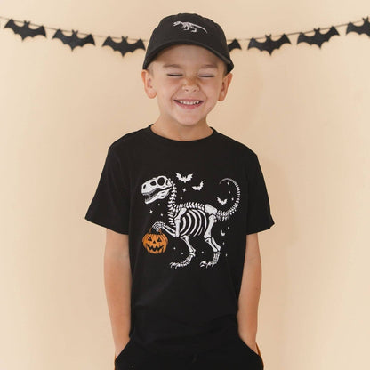 A young boy wearing a black t-shirt with a white skeleton dinosaur and orange pumpkin graphic, paired with a black baseball cap, standing against a beige wall with a bat banner.