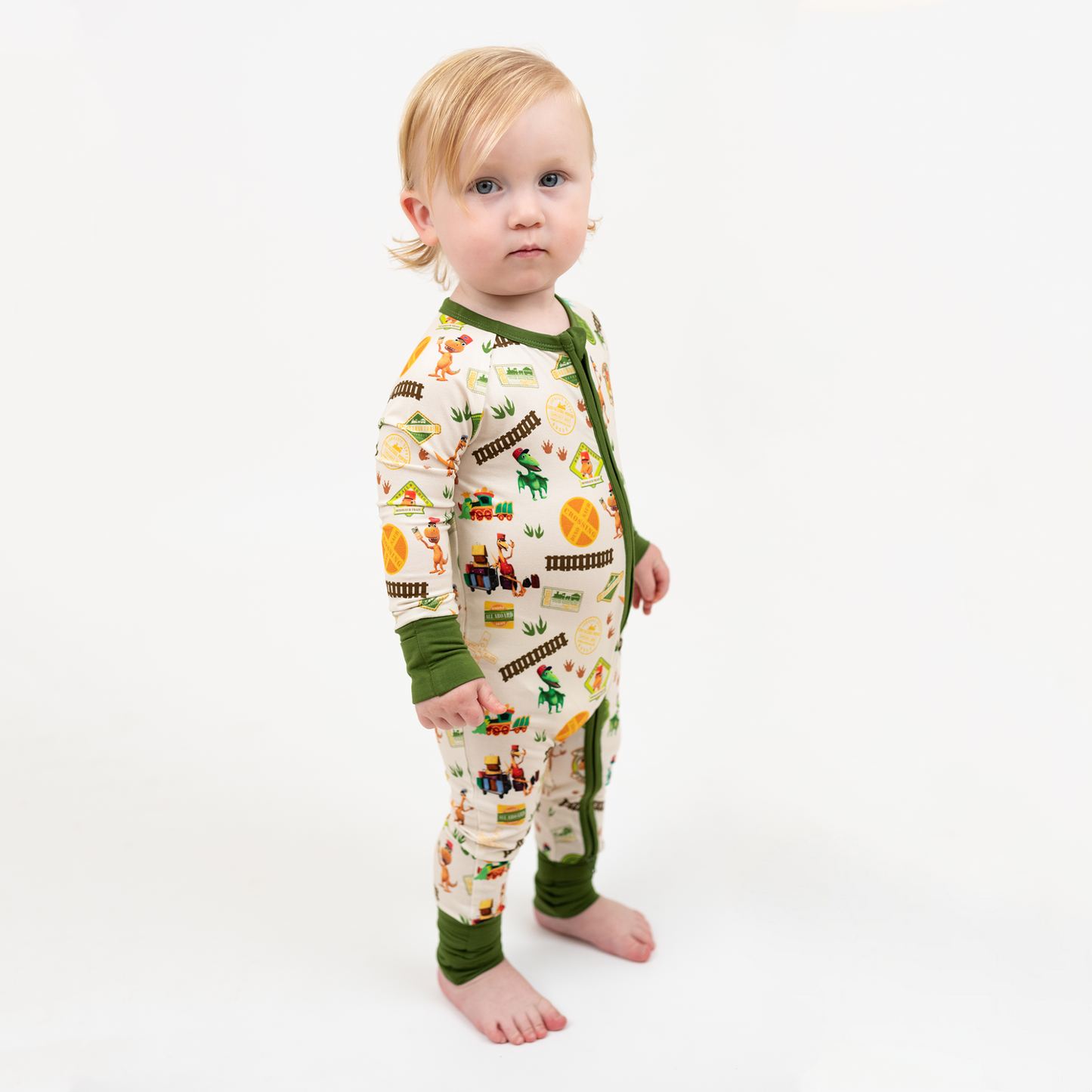 Toddler wearing our Emerson and Friends All Aboard the Dinosaur Train™ bamboo convertible baby pajamas with green trim, double zipper, and colorful orange, blue, and green dinosaurs on train tracks with "Dino Train" print.