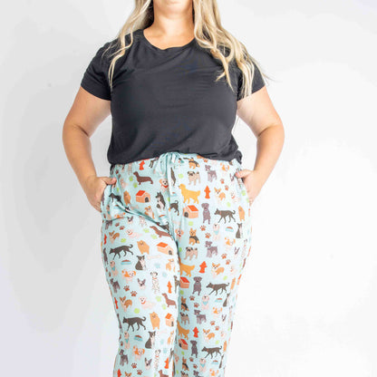 woman wears the "dogs bark" relaxed pants. she rest her hands in the pockets. she wears the solid black tee. the "dogs bark" print is a mix of various types of dogs, dog houses, dog bones, paw prints, dog food, and tennis balls scattered around a light blue background.