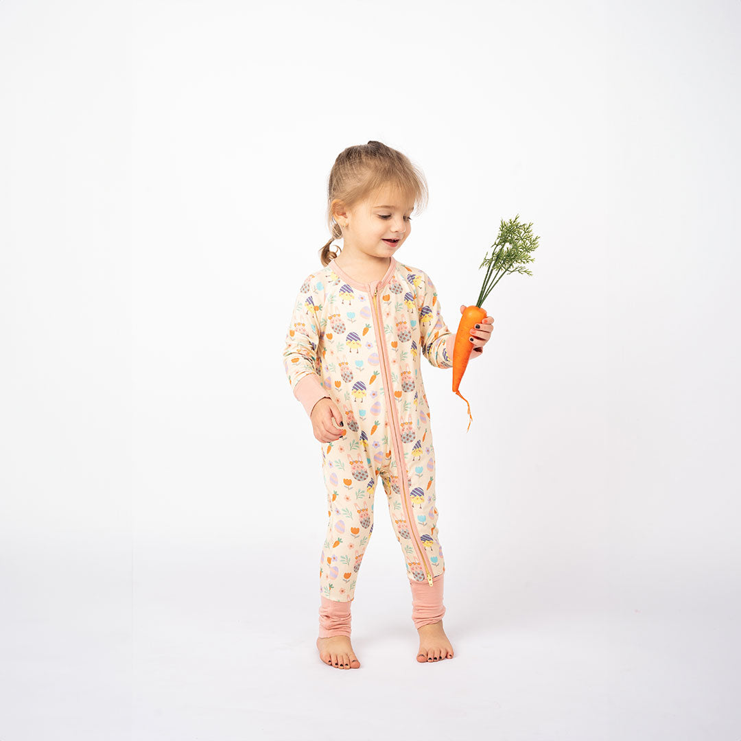 toddler girl holds a large carrot and wears the "egg hunt" convertible. the "egg hunt" print is a easter theme. the colors are pastel. you can find flowers, carrots, easter bunnies in easter eggs, and chicks who have half an easter egg on their head. 