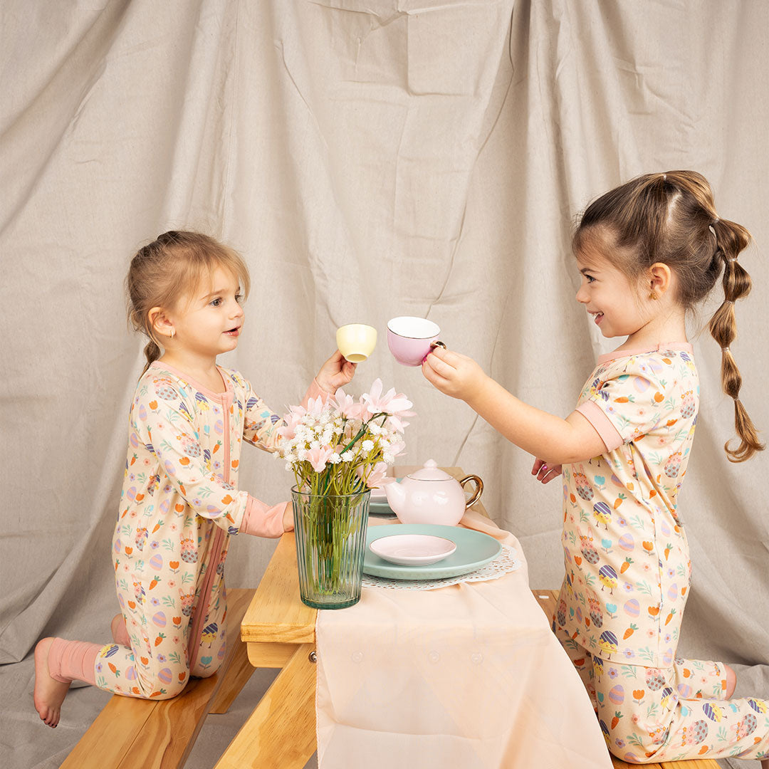 2 girls have a tea party. they sit at a wooden table. there are tea cups, plates, and flowers on the table. one wears the "egg hunt" convertible. the other one wears the "egg hunt" matching pajama set. the "egg hunt" print is a easter theme. the colors are pastel. you can find flowers, carrots, easter bunnies in easter eggs, and chicks who have half an easter egg on their head. 