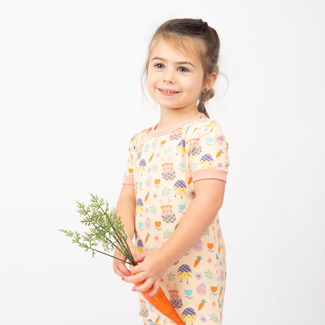 little toddler girl smiles in her "egg hunt" matching pajama set. she holds a large carrot. the "egg hunt" print is a easter theme. the colors are pastel. you can find flowers, carrots, easter bunnies in easter eggs, and chicks who have half an easter egg on their head. 